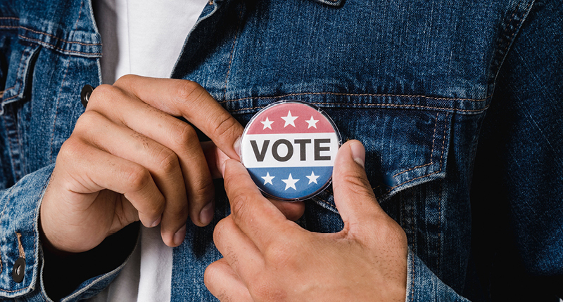 A close photo of a person wearing a jean jacket and pinning a button to the jacket. The button has text that says "vote" with a red stripe and white stars above it and a blue stripe with white stars below it.